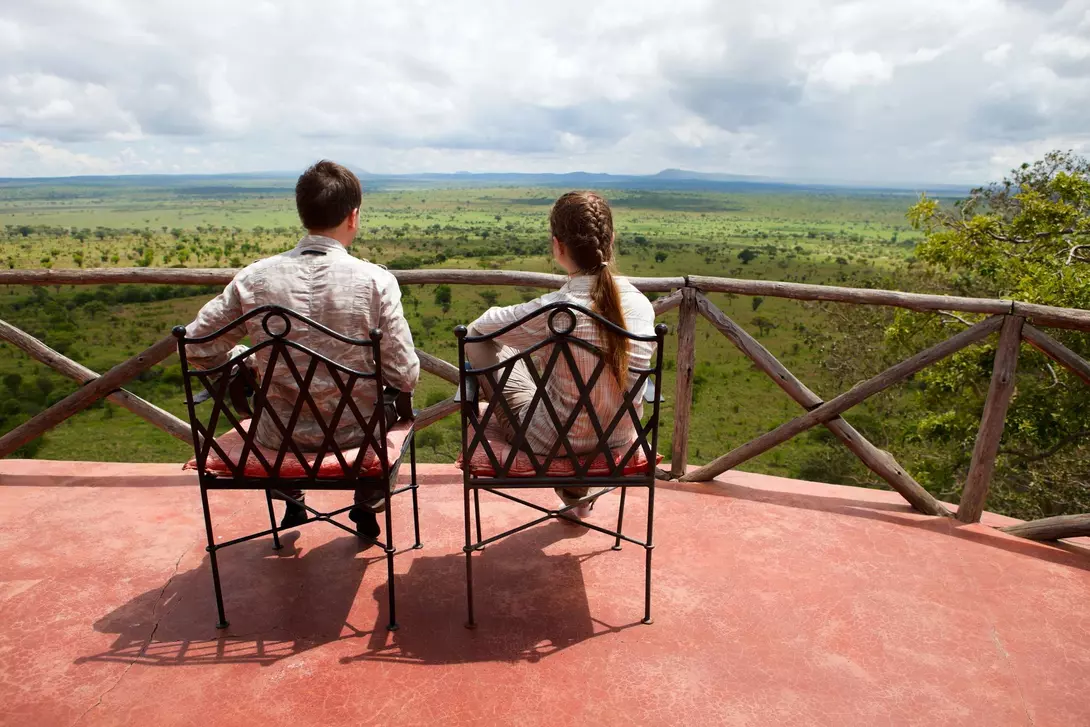 A couple sits in chairs on a balcony, gazing at a lush green landscape under a cloudy sky.