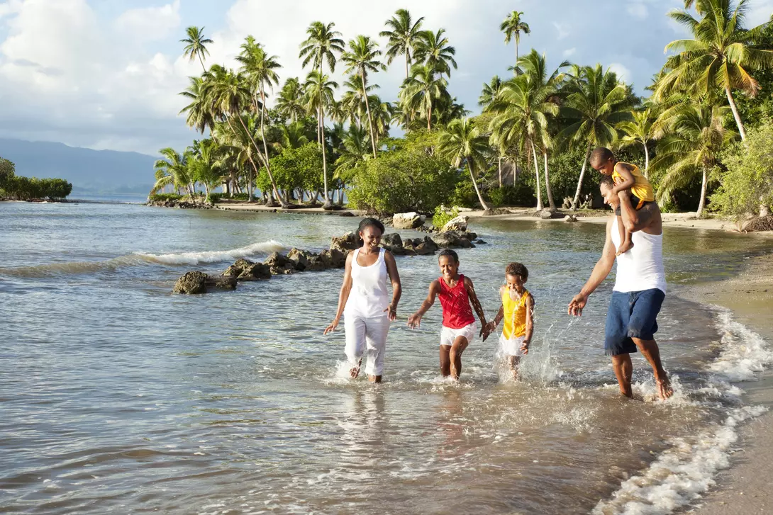 A family with two adults and three children walking in shallow water on a beach, surrounded by palm trees and a scenic backdrop.