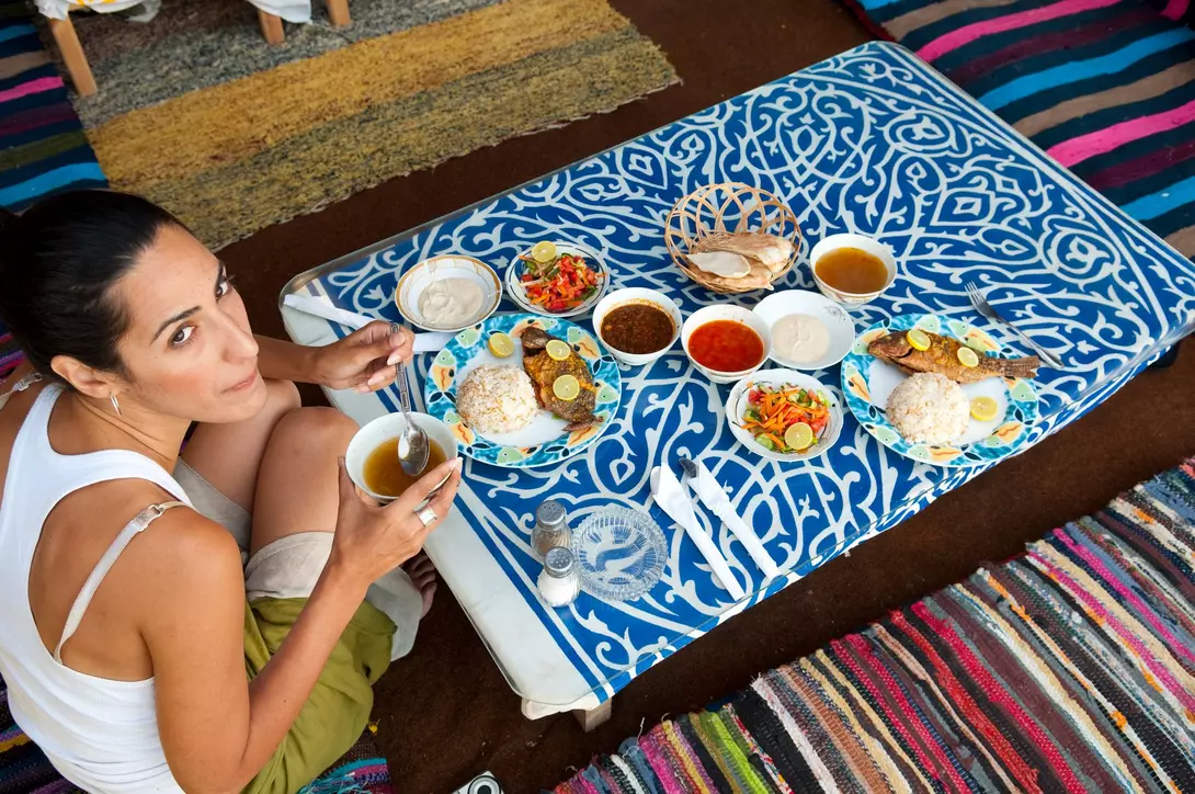 Person sitting on a colorful rug, enjoying a meal with plates of rice, fish, and various side dishes.
