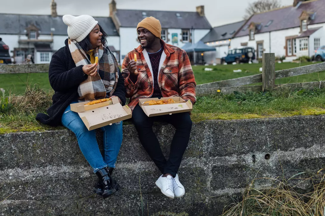 Two people enjoy food from takeout boxes while sitting on a stone wall, with buildings and greenery in the background.