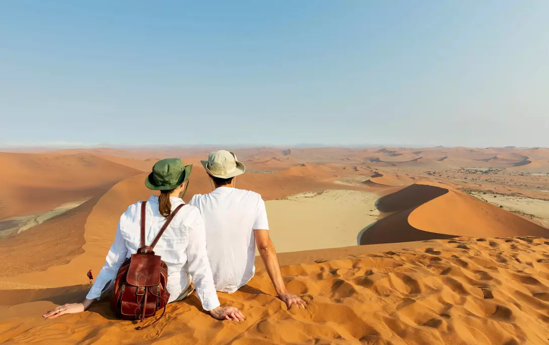A couple sits on a sandy dune, overlooking vast desert landscapes under a clear sky.