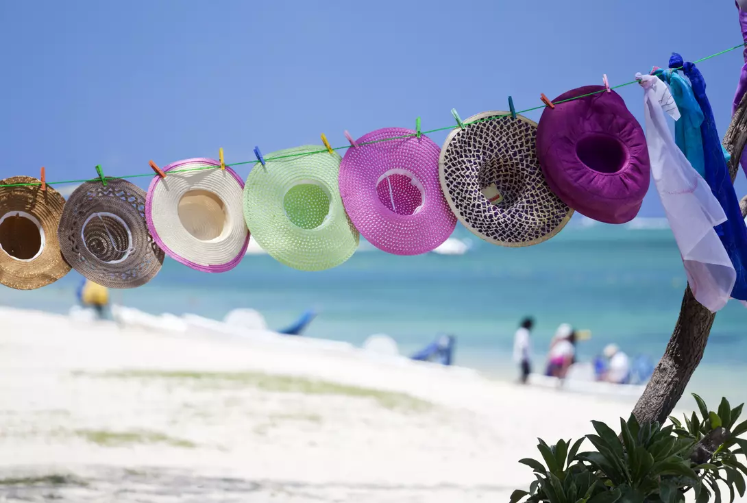 Colorful hats hang on a line with a beach and ocean in the background, accompanied by people enjoying the scenery.