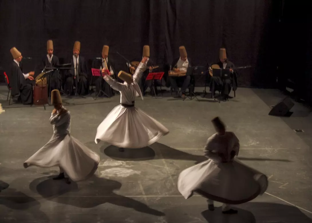 Whirling dervishes perform in front of musicians, all wearing traditional hats, on a dimly lit stage.