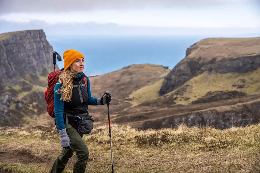A woman with long hair in a bright orange hat hikes along a coastal path, with cliffs and the ocean in the background.