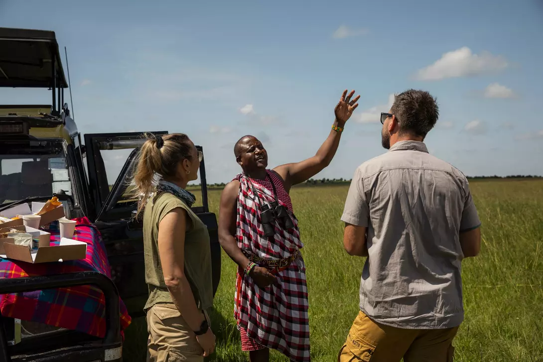 Three people engage in conversation outdoors near a vehicle, surrounded by a grassy landscape under a blue sky.