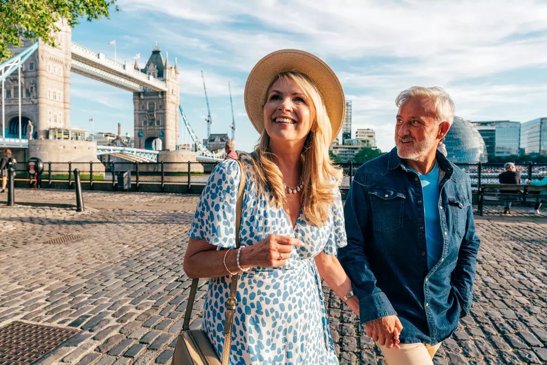 A smiling couple strolls hand in hand near the Tower Bridge in London on a sunny day.