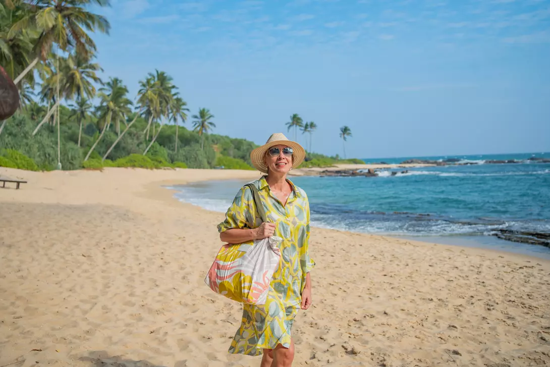 A smiling woman in a yellow dress and hat walks along a sandy beach with palm trees and ocean in the background.
