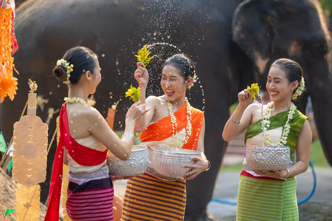 Three women in colorful traditional outfits joyfully splash water while celebrating with an elephant in the background.