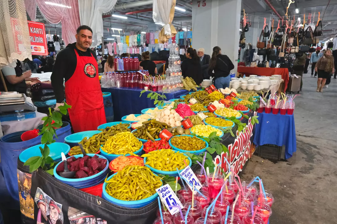 A vendor in an apron stands behind a vibrant table filled with colorful fruits, vegetables, and drinks at a market.