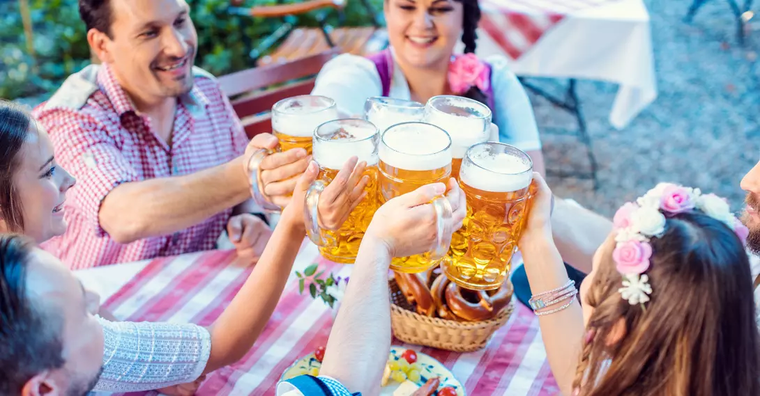 Group of people cheerfully toasting with large beers at a outdoor table, surrounded by pretzels and a festive atmosphere.