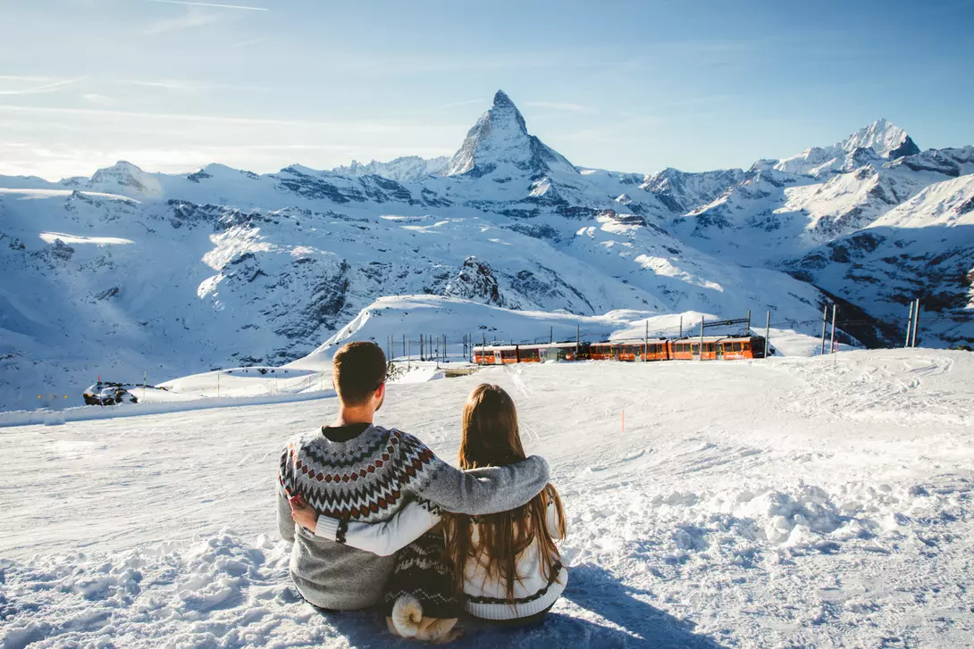 A couple sits together on a snowy slope, overlooking mountains, with the Matterhorn peak in the background.