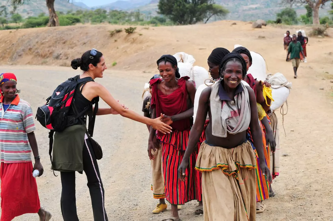 A person greets a group of women in colorful clothing on a dirt road, with mountains in the background.