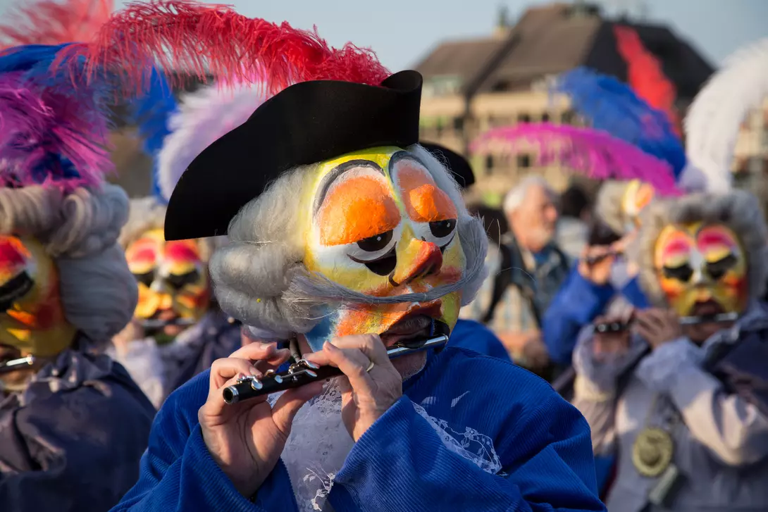 A performer in a feathered hat plays a flute, surrounded by others in colorful masks and costumes during a celebration.