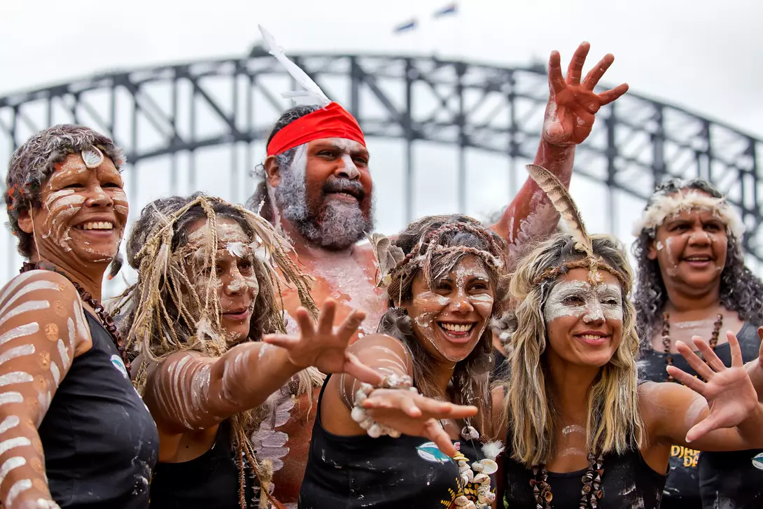 A group of six Indigenous people celebrating with traditional attire and body paint in front of a bridge.