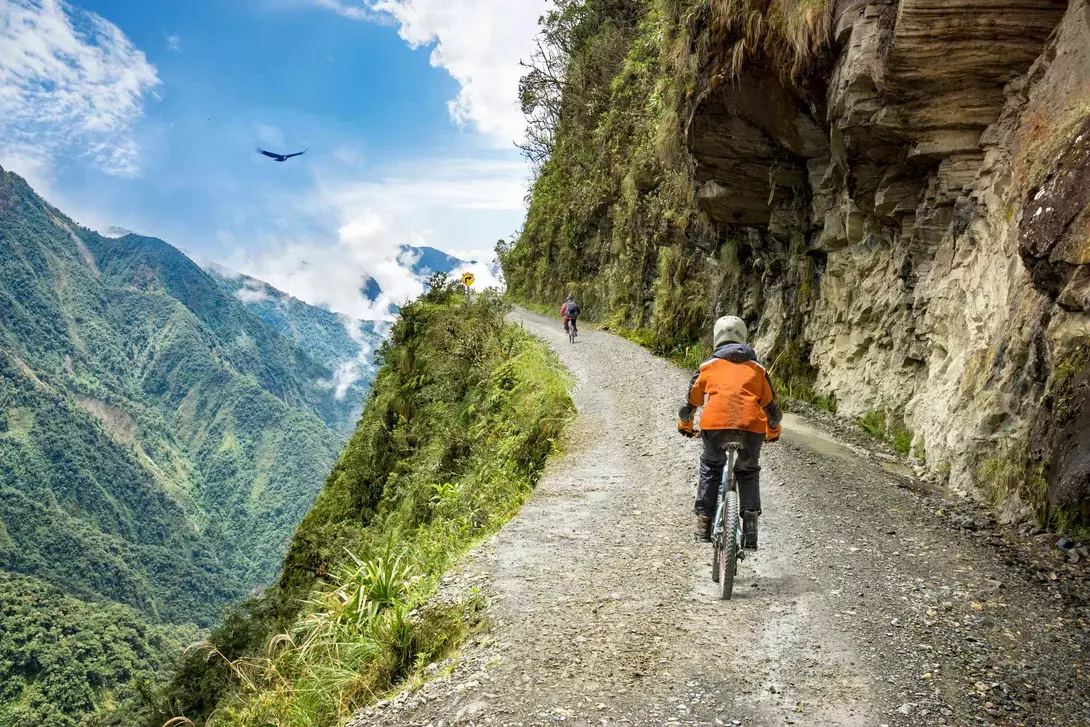 Bike tourists ride on North Yungas road, known as "Death Road" in Bolivia. In the background sky circles a condor over the scene.