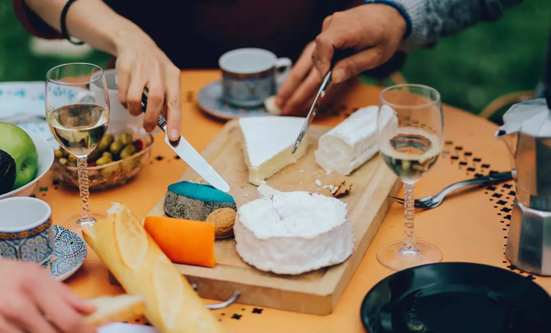 A close-up of a cheese platter with various cheeses, wine glasses, and fruits on a table. Hands are cutting cheese.