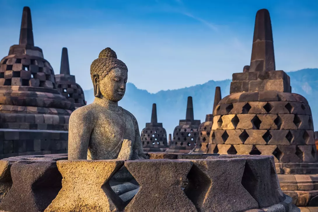 Buddha statue in the foreground with ancient, tiered stupas against a hazy mountain backdrop at sunrise.