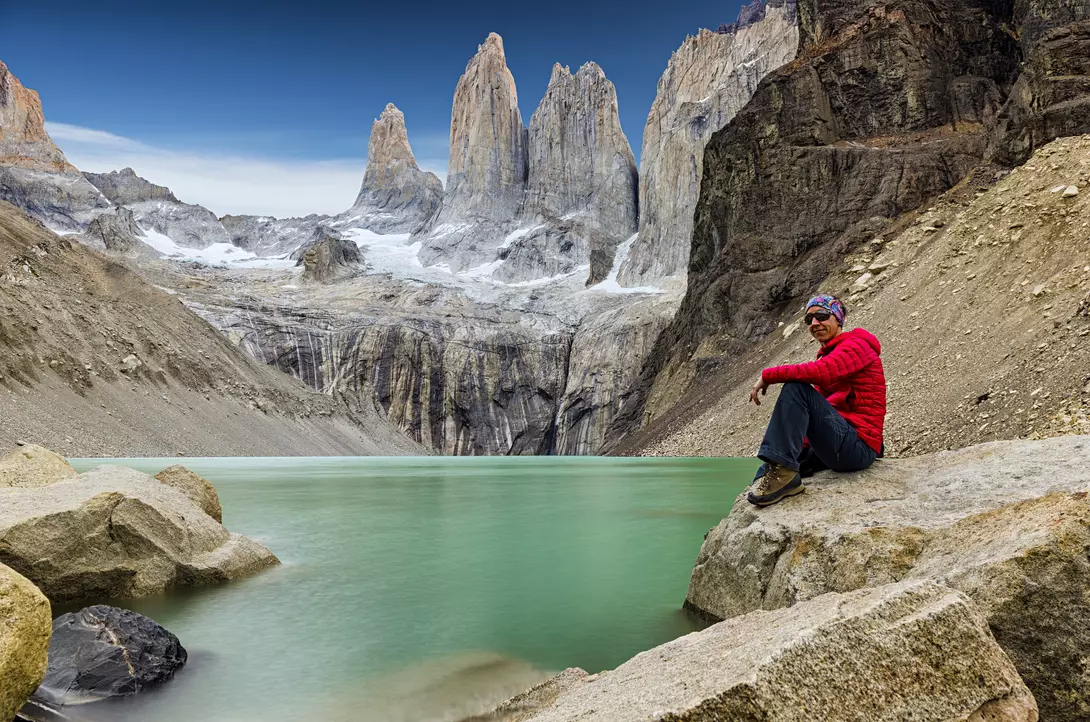 Person in red sitting on rocks by a turquoise lake, with towering mountains in the background under a clear blue sky.