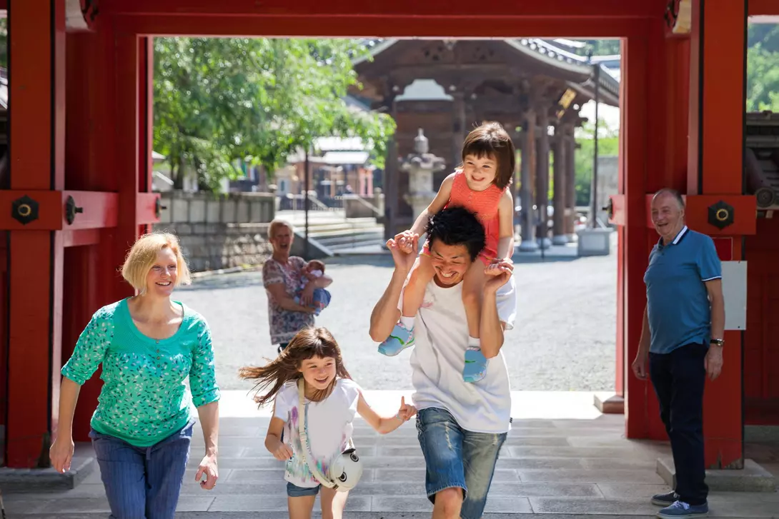 A joyful family walks through a gateway, with a dad carrying a girl on his shoulders and two children running alongside.