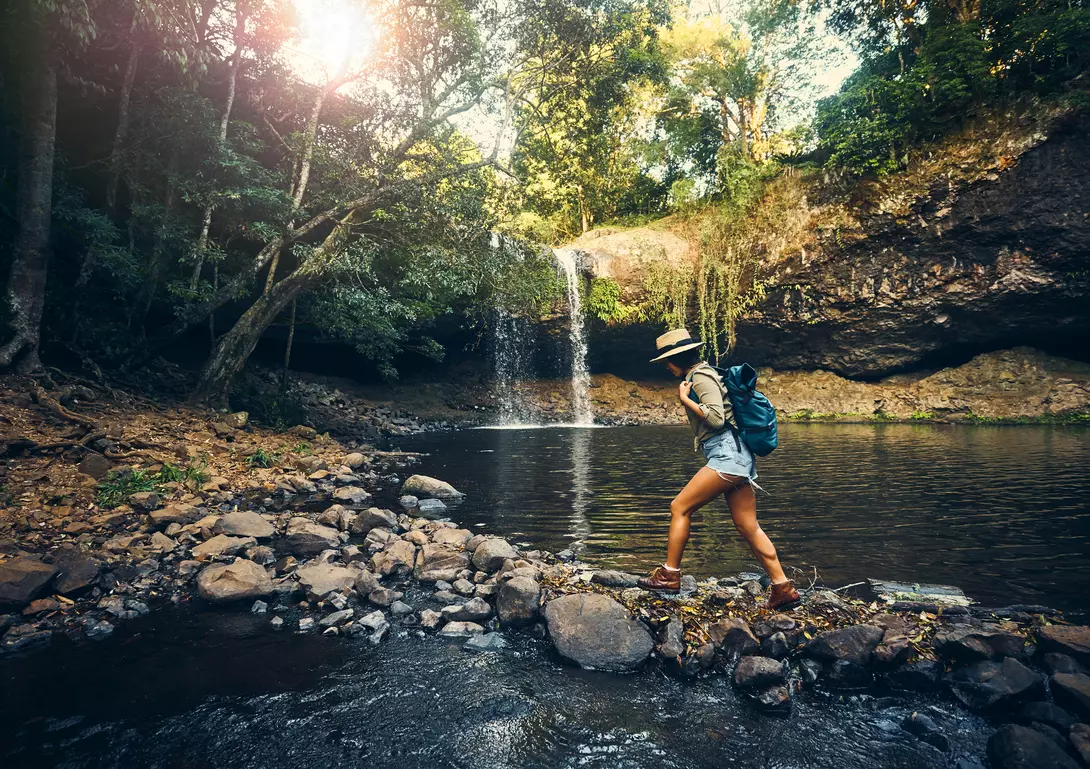 A person in shorts and a hat walks along rocks by a forest stream, with a waterfall in the background.