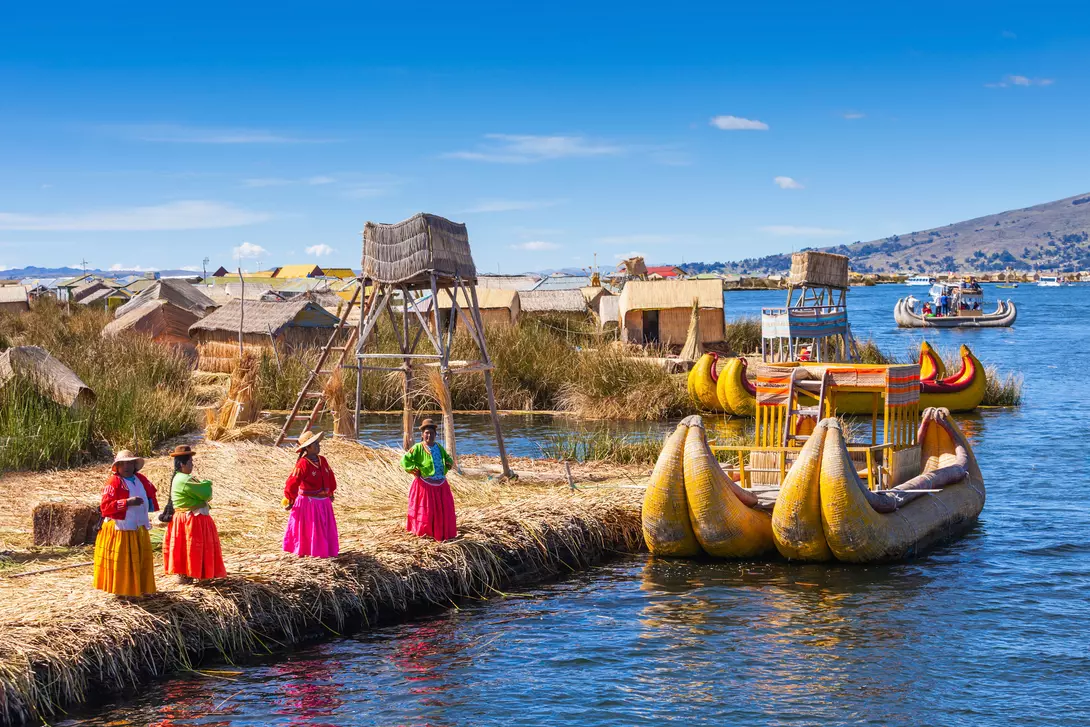 Colorful traditional boats and local women in vibrant clothing by the floating islands on a lake under a clear blue sky.