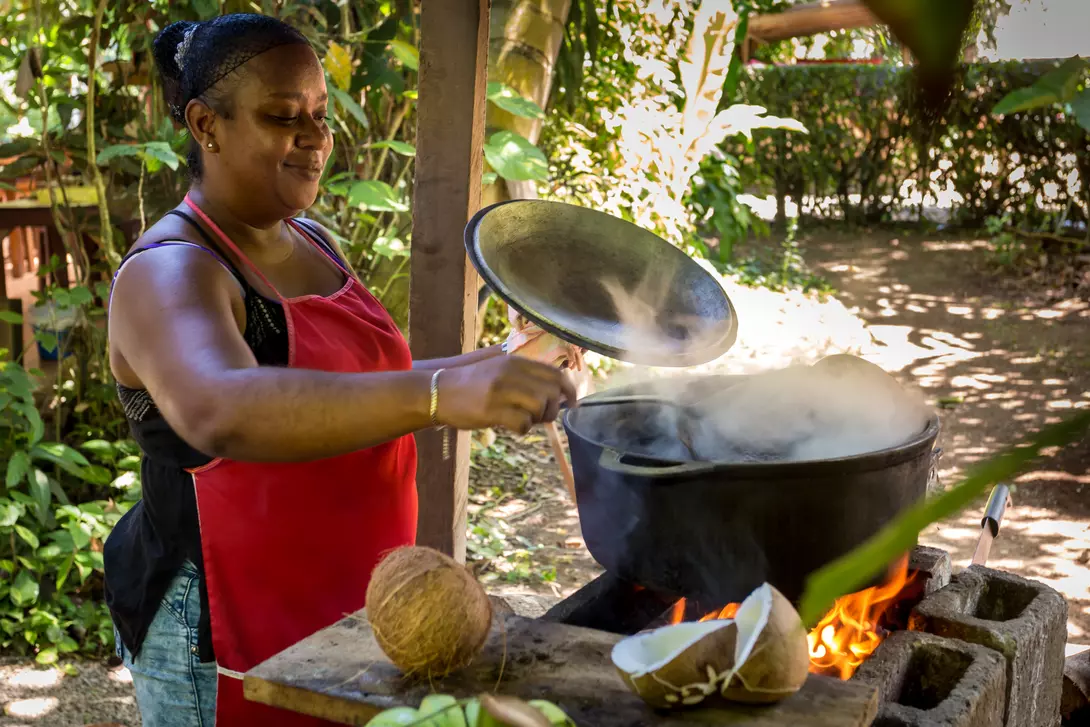 A woman in a red apron stirs a pot over a fire outdoors, surrounded by greenery and coconuts.