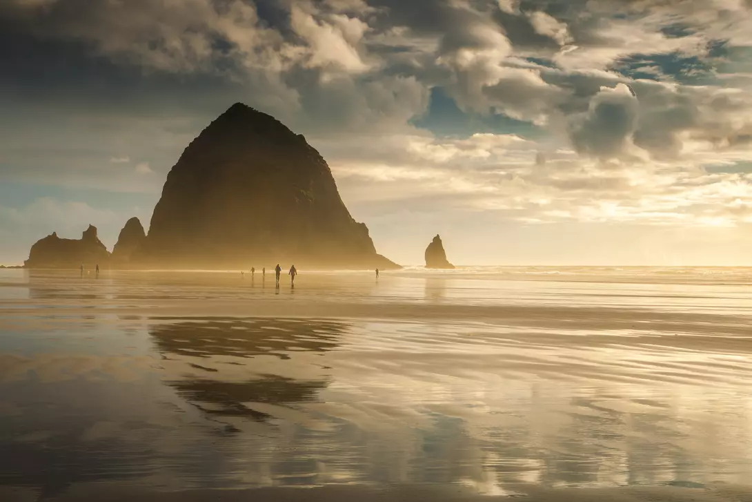 Oregon's Cannon Beach at sunset and in between storms. People can be seen strolling on the beach.