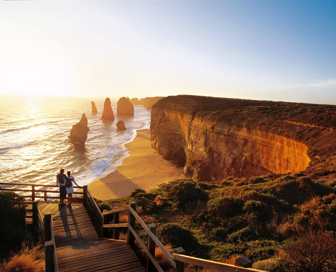 A couple stands on a wooden walkway overlooking a beach with cliffs and sea stacks at sunset.