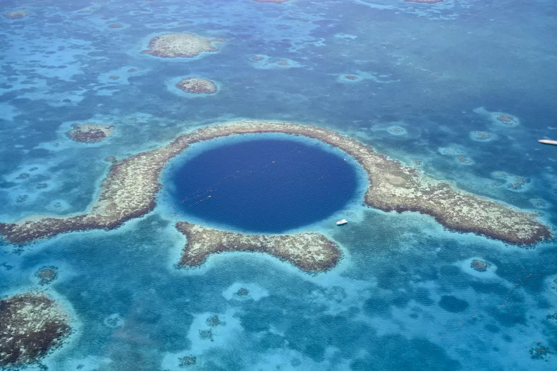 Aerial view of Great Blue Hole a marine sinkhole and geological wonder in the Belize Barrier Reef Reserve. A small white yacht is moored at it's edge