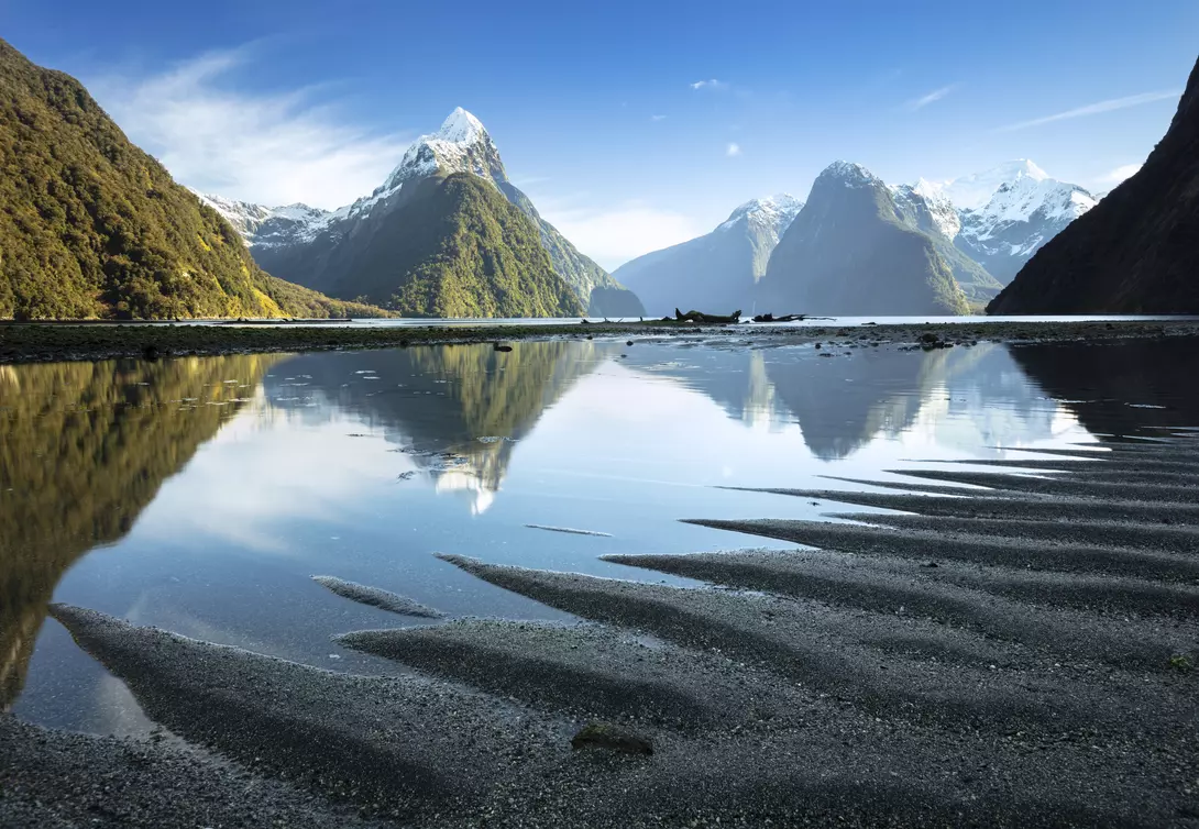 Ripples in the sand point to the tall peaks of Milford Sound which are reflected in the still waters of the fiord