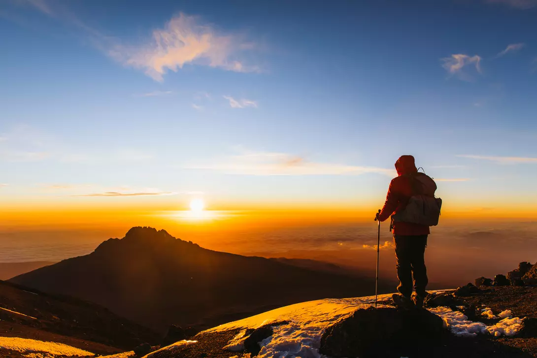 Man with backpack and hiking poles got to the top of Africa - Mount Kilimanjaro and looking at the beautiful bright sunny sunrise