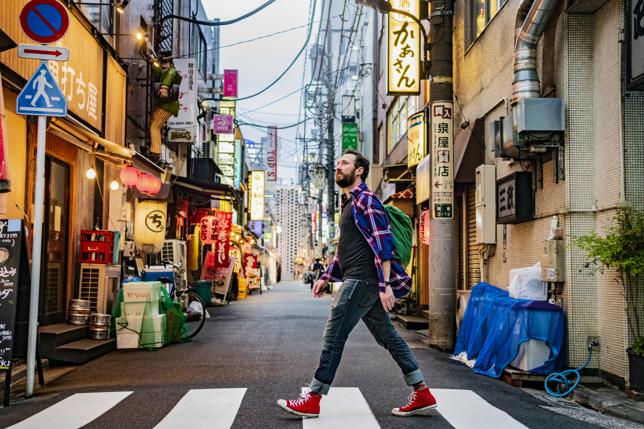 A man in a plaid shirt and red sneakers walks across a crosswalk in a vibrant city street filled with neon signs.